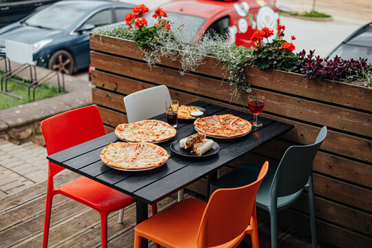 Several pizzas, a grilled wrap, fries and drinks arranged on a colorful outdoor restaurant terrace table beside flower planters