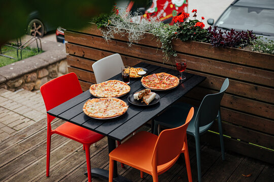 Several pizzas, a grilled wrap, fries and drinks arranged on a colorful outdoor restaurant terrace table beside flower planters