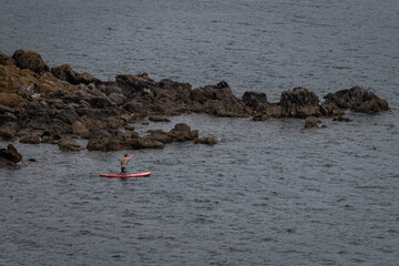 A person kneels on a red stand-up paddle board, paddling near a rugged, rocky shore in open waters. © Sorin