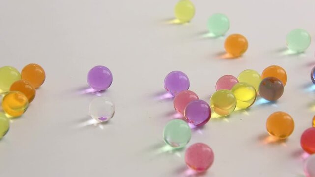 Colorful water gel beads lie scattered across a plain white table, arranged in a loose cluster with orange, purple, green and yellow colors, remaining still in soft daylight.