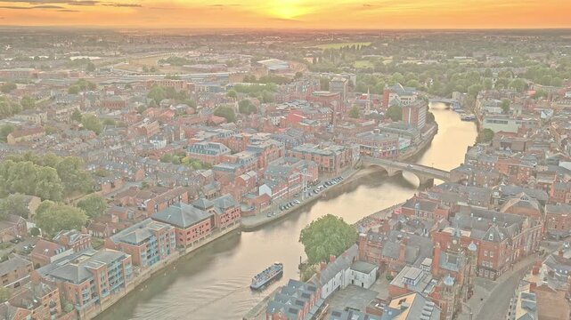 River Ouse winds through York, England, beneath Skeldergate Bridge, with King's Staith, Queen's Staith and dense historic buildings glowing in sunset light, drone forward flyover.