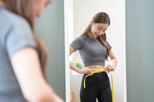 Weight lose, loss concept. Slim body asian young woman hand use tape measuring around waistline in fit sports.