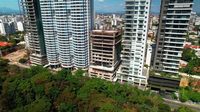 High-angle aerial drone shot flying forward and tracking right parallel to luxury residential skyscrapers and building construction along a tree-lined avenue