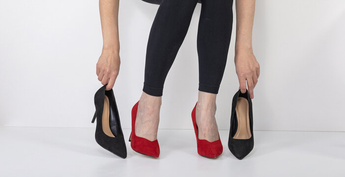 Woman sitting and holding pair of black high heel shoes while wearing red high heel shoes. Choice of footwear for fashion and style. Leg health and comfort concept for tired feet