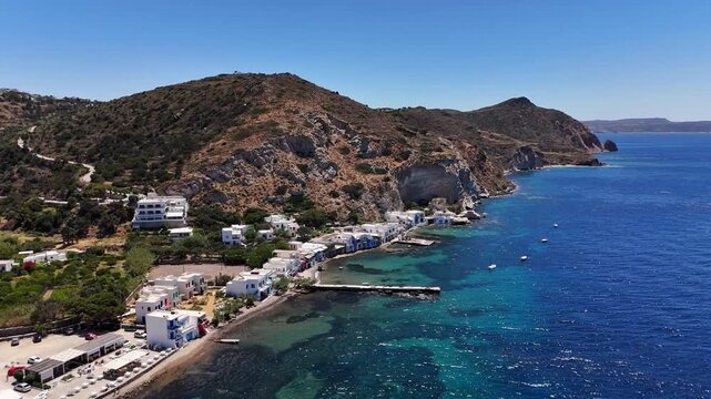 Klima fishing village and colorful syrmata boathouses stretch along a rugged volcanic coastline beside clear Aegean waters, Milos, Greece, drone pullback.