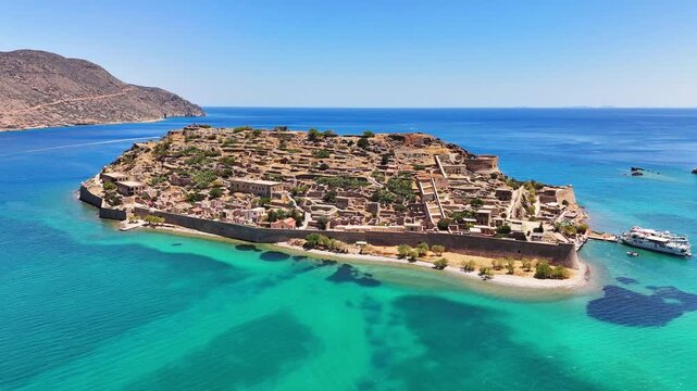 Kalydon, known as Spinalonga, shows Venetian fortress walls and historic ruins across a rocky island surrounded by turquoise waters in Elounda Bay, Crete, Greece, drone pullback.