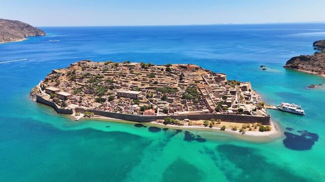 Kalydon Island and Spinalonga fortress show Venetian walls, stone ruins and a harbor with passenger boats above turquoise Mirabello Bay in the Cretan Sea, Lasithi, Greece, drone orbit.