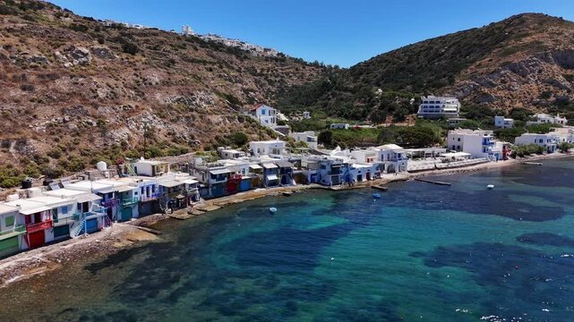 Klima fishing village lines a rocky waterfront with colorful syrmata boathouses beneath dry volcanic hills and clear Aegean water, Milos, Greece, oblique Drone Forward Flyover.
