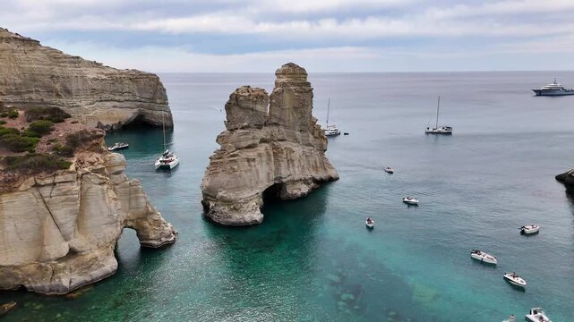 Kleftiko landmark sea stack rises between white volcanic cliffs and sea caves, with motorboats and sailboats anchored in turquoise Aegean Sea waters, Milos, Greece, parallax orbit descent.
