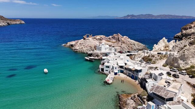 Firopotamos fishing village features white syrmata boat houses, Agios Nikolaos chapel and rocky volcanic cliffs around a turquoise cove on the Aegean Sea, Milos, Greece, drone forward flyover.