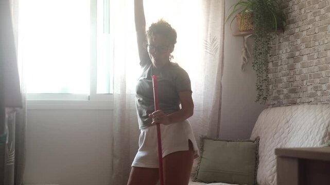 Joyful middle-aged woman having fun while doing chores, dancing and singing with a broom in her living room, enjoying her domestic work and feeling happy with a positive attitude