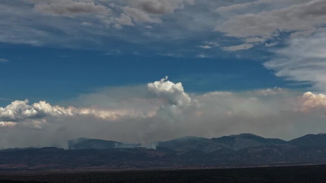 4K aerial timelapse of a single wildfire smoke plume billowing upward near Mt Baldy in New Mexico. A distinct pyrocumulus column rises above the mountain ridge into a partly cloudy blue sky.