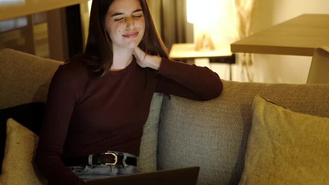 Tired woman working on her laptop late at night at home feeling neck pain and discomfort from overwork, massaging her stiff muscles with her hand while sitting on the couch in the dark