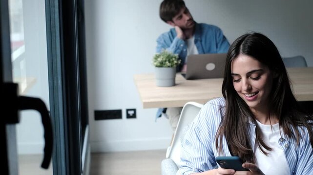 Beautiful young woman smiling while texting on her smartphone at home, with a jealous and curious man in the background looking at her while he's working on his laptop computer