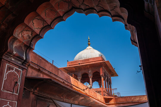 White marble dome chhatri framed by ornate red sandstone scalloped arch against vibrant clear blue sky at Jama Masjid, Delhi, India.