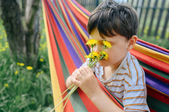 Child smelling a yellow dandelion in a colorful hammock outdoors