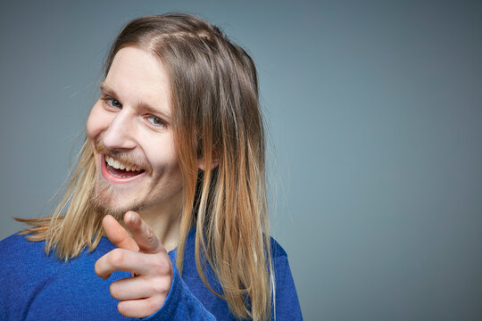 Portrait of laughing young man with long blond hair pointing on viewer