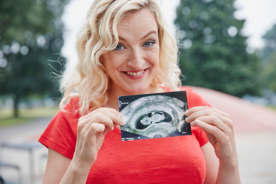 Portrait of happy pregnant woman holding ultrasound image outdoors