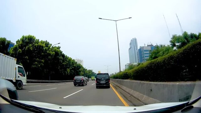 Car dashboard camera point of view driving on urban highway toll road past delivery cargo truck and tall modern skyscraper buildings in metropolitan city during daylight traffic commute footage