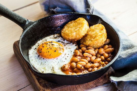 Fried egg, baked beans and hash browns in frying pan on wooden board