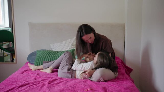 A cheerful mother and daughter play, laugh, and cuddle together while lying on a bed in a bright bedroom.