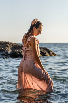Golden hour coastal scene with adult female standing in ocean water wearing wet pink dress
