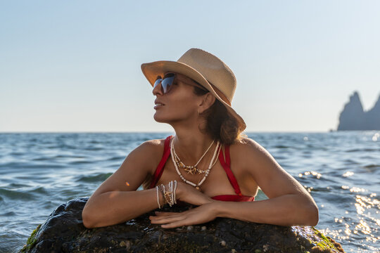 Sunbathing vacation featuring mature woman wearing straw hat and sunglasses leaning on rock in sea
