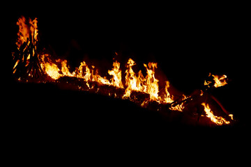 A horizontal trail of bright orange flames and burning wood logs glowing intensely against a pitch-black background, creating a dramatic and fiery night scene © Syarif