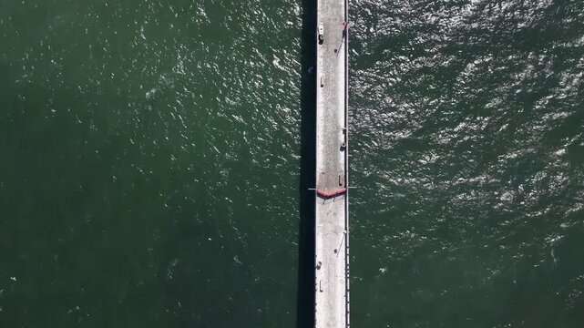 Top-down aerial drone view of the Pacifica Municipal Pier in California, showing people walking along the concrete structure over the deep green, sun-dappled waters of the Pacific Ocean