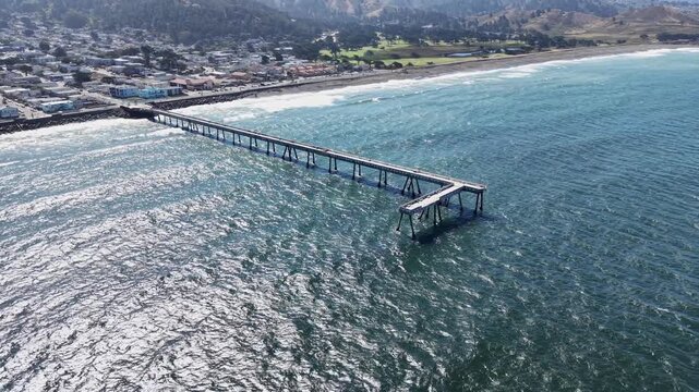 Morning drone video captures Pacifica Pier stretching into shimmering blue ocean, dramatic coastline, rolling waves, and sunlit seaside town. Vibrant natural light, wide aerial footage.