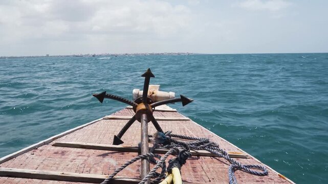 Front view of metal boat anchor resting on wooden deck of vessel sailing across deep blue ocean towards distant coastline.