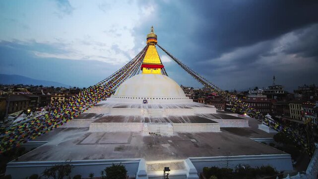 Clouds drift over the Boudhanath Stupa and its golden spire as prayer flags sway and crowds gather below during the transition from dusk to night, Time Lapse