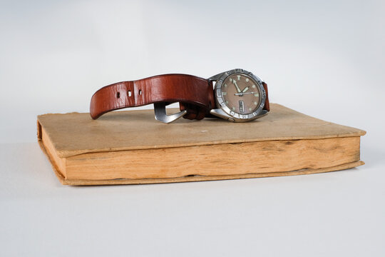 Studio shot of a classic analog watch with a brown leather strap resting on an old vintage book, isolated on a white background.