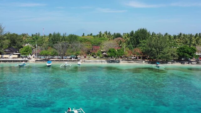 Aerial drone flies along the Gili Trawangan coast facing the island, over clear blue-green shallow water with the sandy bottom visible, boats, beach huts, lounge chairs and trees, Lombok, Indonesia.