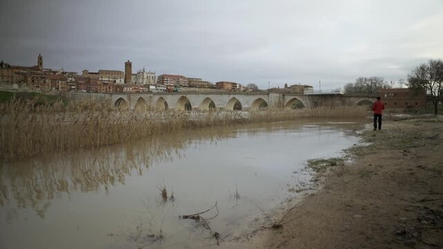 Man in Red Jacket Walking Along Flooded Muddy Riverbank in Winter