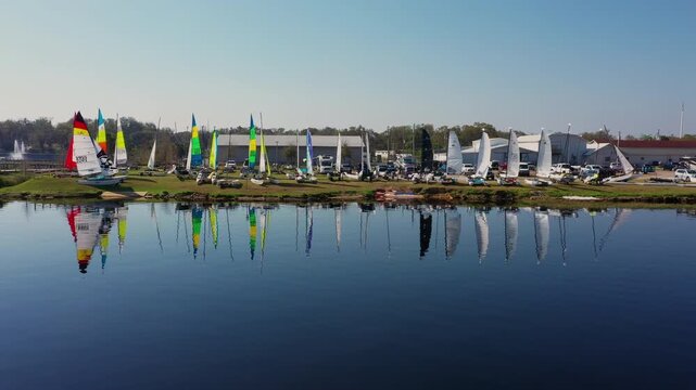 Aerial view of a lively sailing regatta on the shores of Lake Minneola in Clermont, Florida, with rows of colorful catamarans, parked cars, and lakeside clubhouses.