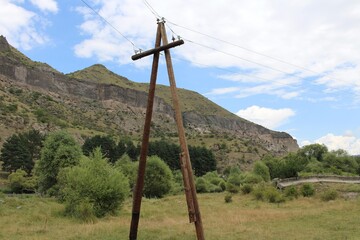 A-frame wooden power pole stands in a green valley with the historic Vardzia cave monastery complex and rocky cliffs visible in the background. © StasiaVisualStock