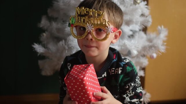Christmas celebration. Child wearing funny festive eyeglasses holding Christmas present in his hands at home