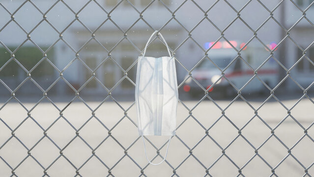 Medical mask hanging on chain link fence near blurred ambulance, virus prevention and healthcare emergency