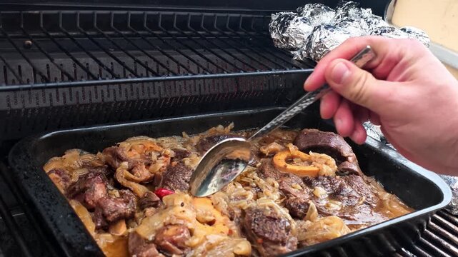 Hand stirs braised meat in pan over grill. Steam rises from tender beef, onions, and sauce while foil wrapped potatoes warm above rack in backyard cooking, with spoon moving through hot gravy.