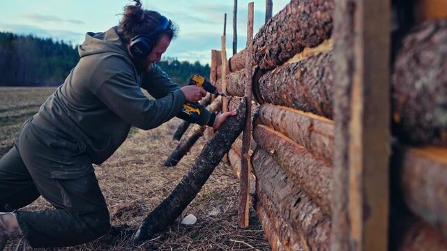 Man using a cordless power drill to secure wooden framing or logs on an outdoor wooden structure during construction work