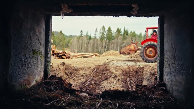 A man operates a red agricultural tractor with a front-end loader in a wooded forestry area in Norway