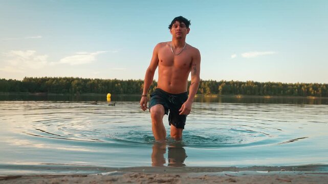 Young hispanic man walking out of the lake water to the beach during golden hour, active guy in nature landscape.