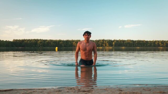 Front shot of a shirtless young Hispanic male in swim trunks walking out of a calm lake onto the sandy shoreline at golden hour.