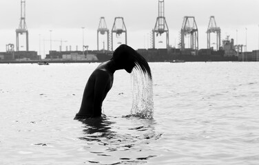 Fototapeta premium Silhouette of a young man flipping long hair back in sea water, port cranes background