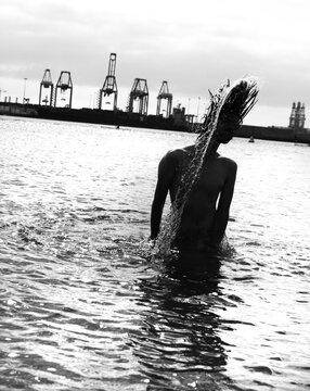 Silhouette of a young man flipping long hair back in sea water, port cranes background