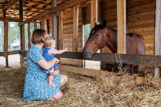 A mother and child crouched on hay inside a wooden barn looking at a horse