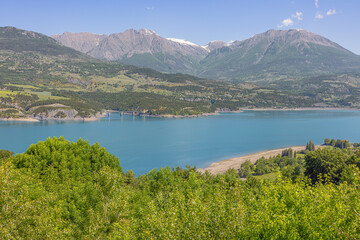 Obraz premium Snowcapped mountains above the Lac de Serre Poncon with the bridge over the Riou-Bourdu river, seen from the south shore