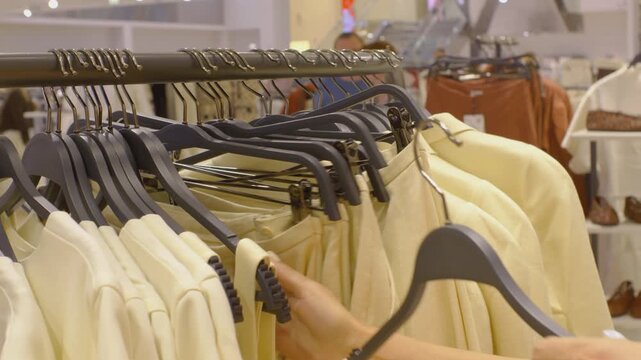 A woman examines clothes on a hanger in a modern, brightly lit boutique, carefully feeling the cream and pastel fabrics. A close-up of her manicured hand is shown as she pulls out a hanger to assess t