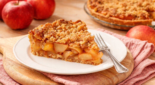 Fresh apple pie slice is served on a plate next to whole apples and a pie on a wooden table
